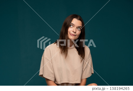 Young woman making a goofy facial expression with tongue out, looking sideways, playful and humorous on teal background 138194265