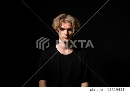 Young blond man with beard looking directly at camera, standing in a studio with a dark background, portraying seriousness and introspection Young blond man with beard looking directly at camera, standing in a studio with a dark background, portraying seriousness and introspection 138194314