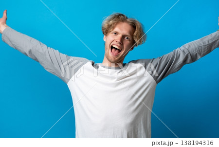 Excited young man with outstretched arms celebrating success, expressing happiness and joy on blue background, feeling triumph and achieving goals 138194316