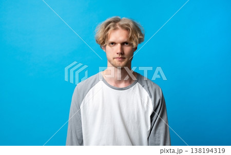 Young casual man with a serious expression and blonde wavy hair in a raglan shirt, standing confidently against a bright blue background, copy space Young casual man with a serious expression and blonde wavy hair in a raglan shirt, standing confidently against a bright blue background, copy space 138194319