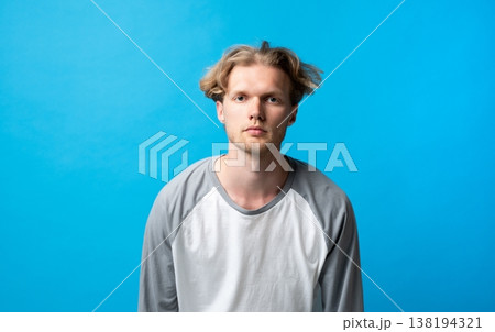 Young man with blond hair and a neutral expression standing directly facing the camera, wearing a casual long-sleeve t-shirt against a striking blue studio backdrop 138194321
