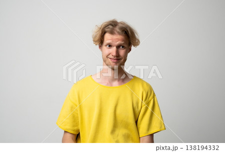 Young blonde man feeling awkward, smirking and glancing sideways, showing skepticism while standing in a yellow t-shirt on a solid grey background 138194332