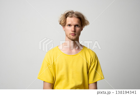 Young man with blond hair and light stubble wearing a casual yellow t-shirt, standing confidently in a studio, presenting a serious and neutral expression while looking directly at the camera 138194341