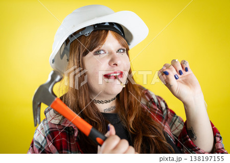 Woman wearing hard hat holding hammer with nail in mouth Woman wearing hard hat holding hammer with nail in mouth 138197155