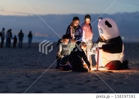 Children and adult in panda costume sharing light on beach at dusk Children and adult in panda costume sharing light on beach at dusk 138197191