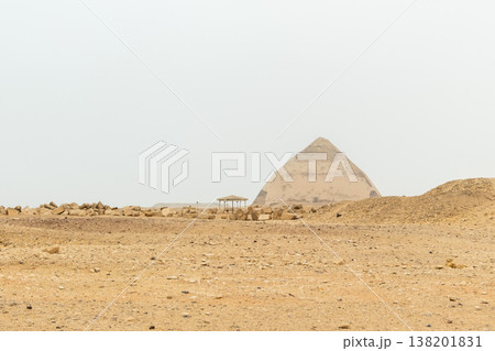 A white pyramid stands in the desert landscape of Dahshur, Egypt. The structure is partially obscured by a hazy sky, surrounded by sandy terrain. 138201831