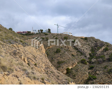 Solitary cyclist traverses rugged hillside road beneath gray sky with distant cranes and houses 138203135