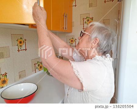 An elderly woman wearing glasses and a white blouse opens a yellow wall cabinet in the kitchen. An elderly woman wearing glasses and a white blouse opens a yellow wall cabinet in the kitchen. 138205504
