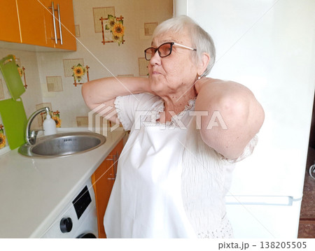 An elderly woman wearing glasses and a white apron stands in the kitchen. 138205505