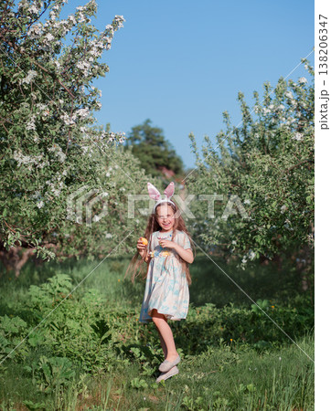Cute girl wearing bunny ears holding colorful Easter eggs over eyes standing near blooming tree Cute girl wearing bunny ears holding colorful Easter eggs over eyes standing near blooming tree 138206347