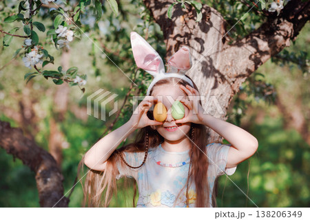 Cute girl wearing bunny ears holding colorful Easter eggs over eyes standing near blooming tree Cute girl wearing bunny ears holding colorful Easter eggs over eyes standing near blooming tree 138206349