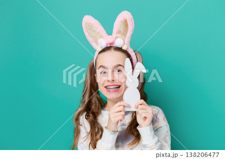 Teenage girl with bunny ears holding wooden bunny toys smiling on bright green background studio 138206477