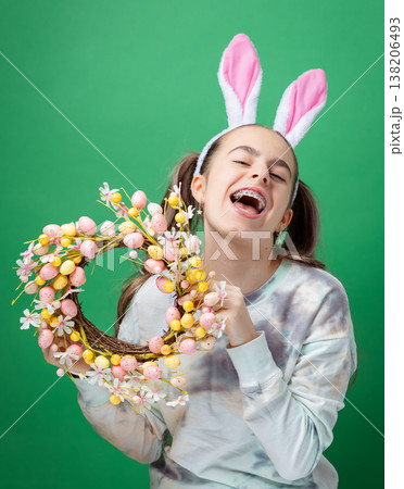 Teenage girl with bunny ears holding decorative Easter wreath made of flowers eggs green background. 138206493