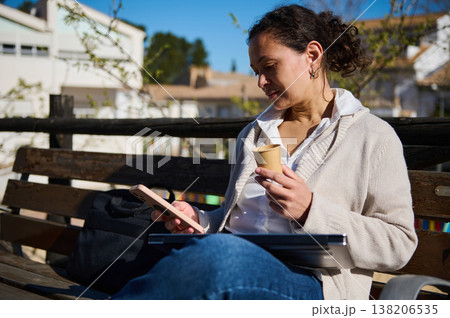 Woman Using Phone and Drinking Coffee While Working Remotely on a Bench Outdoors Woman Using Phone and Drinking Coffee While Working Remotely on a Bench Outdoors 138206535