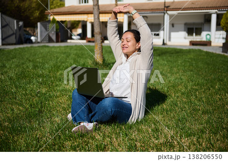 Woman Working On Laptop Outdoors Sitting On Grass Stretching During Remote Work Break In Suburban Yard 138206550