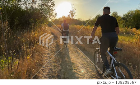 Young male friends ride mountain ski bikes along trail on sunny summer day. Beautiful golden sunlight at background. Concept of active lifestyle, adventure spirit and carefree weekend outdoor 138208367