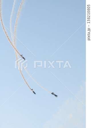 Three aerobatic aircraft performing a synchronized aerial maneuver with curved smoke trails against a clear blue sky during an aviation airshow. Dynamic formation display in an open sky background Three aerobatic aircraft performing a synchronized aerial maneuver with curved smoke trails against a clear blue sky during an aviation airshow. Dynamic formation display in an open sky background 138210805