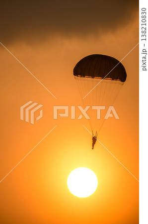 Combat paratrooper deploying a parachute and descending through a glowing orange sunset sky below dark clouds. Silhouette paratrooper during an aerial demonstration Combat paratrooper deploying a parachute and descending through a glowing orange sunset sky below dark clouds. Silhouette paratrooper during an aerial demonstration 138210830