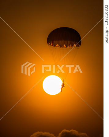 Silhouette of a parachutist descending in front of a glowing sun. A combat paratrooper with a warm orange sky during an airshow demonstration 138210832