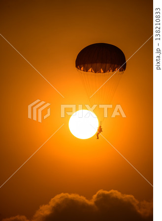 Silhouette of a parachutist descending in front of a glowing sun. A combat paratrooper with a warm orange sky during an airshow demonstration Silhouette of a parachutist descending in front of a glowing sun. A combat paratrooper with a warm orange sky during an airshow demonstration 138210833