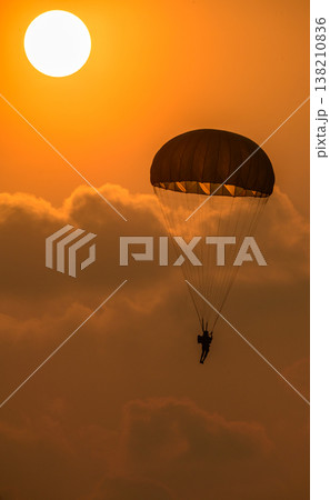 Paratrooper descending in front of a glowing sun. Silhouette of a parachutist against a warm orange sky during an airshow demonstration Paratrooper descending in front of a glowing sun. Silhouette of a parachutist against a warm orange sky during an airshow demonstration 138210836