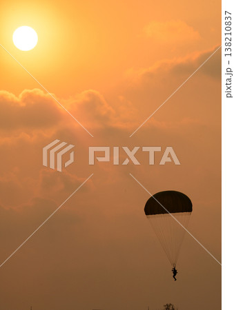 Paratrooper descending in front of a glowing sun. Silhouette of a parachutist against a warm orange sky during an airshow demonstration Paratrooper descending in front of a glowing sun. Silhouette of a parachutist against a warm orange sky during an airshow demonstration 138210837