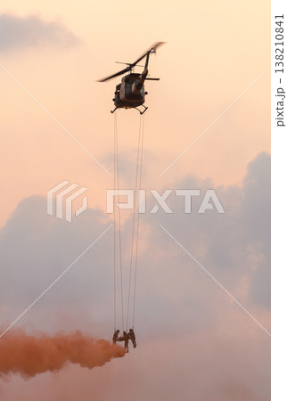 Medics airlift an injured soldier beneath a hovering helicopter using ropes and smoke cover during a rescue demonstration against a sunset sky with clouds at an air show event 138210841