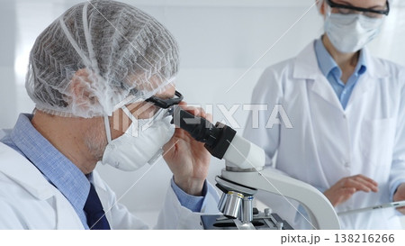 A male scientist wearing a protective mask, glasses and hairnet works with a microscope while a female colleague helps him by holding a tablet and taking notes in a sterile laboratory. Concept of A male scientist wearing a protective mask, glasses and hairnet works with a microscope while a female colleague helps him by holding a tablet and taking notes in a sterile laboratory. Concept of 138216266