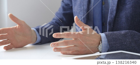 Professional businessman gesturing while discussing strategy, seated at white desk with tablet, eyeglasses, and wristwatch reflecting on sleek workspace surface. Business people concept Professional businessman gesturing while discussing strategy, seated at white desk with tablet, eyeglasses, and wristwatch reflecting on sleek workspace surface. Business people concept 138216340