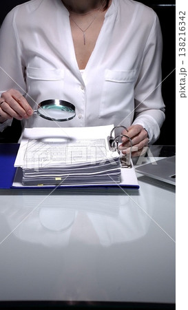 Businesswoman conducting detailed audit checking financial spreadsheets using magnifying glass with laptop and documents on desk. Audit and taxes concept 138216342