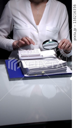 Businesswoman conducting detailed audit checking financial spreadsheets using magnifying glass with laptop, calculator and documents on desk. Audit and taxes 138216356