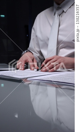 Man in white shirt reviewing legal documents, checking details of a contract with a magnifying glass on a dimly lit desk. Audit, business and overworking concepts 138216357