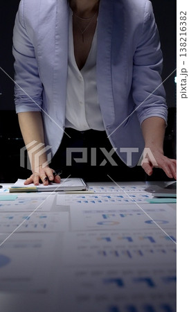 Businesswoman analyzing corporate financial data and sales growth charts on documents spread across an office desk, working late night in a professional setting 138216382