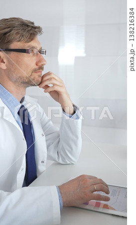 Medical male professional wearing lab coat and glasses is thinking and analyzing data on a tablet, thoughtfully considering information in a bright, modern laboratory setting. Medicine and science 138216384