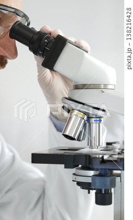 Close up of male researcher examining sample through microscope, wearing safety eyewear in sterile laboratory setting, surrounded by scientific glassware. Medicine, healthcare and science concept Close up of male researcher examining sample through microscope, wearing safety eyewear in sterile laboratory setting, surrounded by scientific glassware. Medicine, healthcare and science concept 138216428