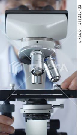 Female scientist wearing lab coat and protective glasses using a modern microscope in a laboratory setting, conducting research and experiments, close up. Health care, medicine and science concept Female scientist wearing lab coat and protective glasses using a modern microscope in a laboratory setting, conducting research and experiments, close up. Health care, medicine and science concept 138216432