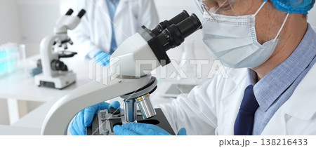 Male scientist in protective mask, cap and gloves studying samples under microscope in sterile laboratory. Concept of medicine, healthcare and science Male scientist in protective mask, cap and gloves studying samples under microscope in sterile laboratory. Concept of medicine, healthcare and science 138216433