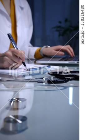 Stethoscope resting on a white reflective desk while medical professional working in clinic office. Wearing lab coat. Typing on laptop and writing notes. 138216480