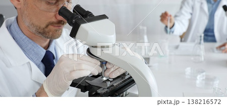 Researcher examining sample through microscope, wearing safety eyewear in sterile laboratory setting, surrounded by scientific glassware. Medicine, healthcare and science concept Researcher examining sample through microscope, wearing safety eyewear in sterile laboratory setting, surrounded by scientific glassware. Medicine, healthcare and science concept 138216572