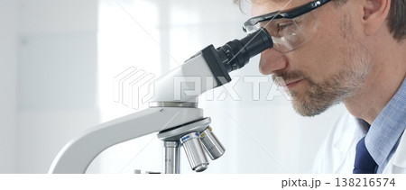 Adult male scientific researcher analyzing sample through microscope, wearing protective gear in high tech laboratory environment. Healthcare, medicine and science concept Adult male scientific researcher analyzing sample through microscope, wearing protective gear in high tech laboratory environment. Healthcare, medicine and science concept 138216574