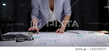 Businesswoman leaning over desk, working late in dark office, analyzing charts and graphs on paperwork, developing financial strategy and preparing for a deadline 138216575