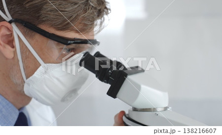 Male scientific researcher wearing protective gear examining sample through microscope, performing detailed laboratory investigation. Medicine and science concept Male scientific researcher wearing protective gear examining sample through microscope, performing detailed laboratory investigation. Medicine and science concept 138216607