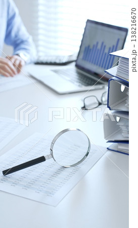 Close up of magnify glass over financial documents near blue folders full of financial papers, accountants are analyzing reports and documents at the background. Audit and taxes in business 138216670