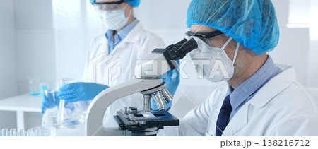 Masked scientist examining specimen under microscope while laboratory assistant handles glassware in a professional medical investigation. Health care and science concept 138216712
