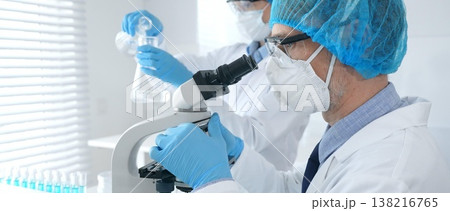 Masked male scientist in protective gear examining specimen under microscope while laboratory assistant handles glassware at the background. Medicine, science concept Masked male scientist in protective gear examining specimen under microscope while laboratory assistant handles glassware at the background. Medicine, science concept 138216765