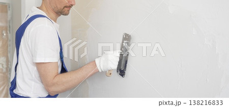 Construction worker plastering white wall, wearing blue overalls and protective white gloves during apartment renovation project 138216833