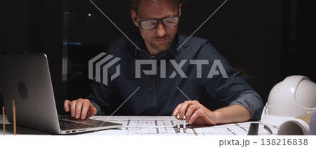 Architect or engineer working late, analyzing building blueprints and plans with a laptop, focusing on detailed architectural design during an intense night shift 138216838