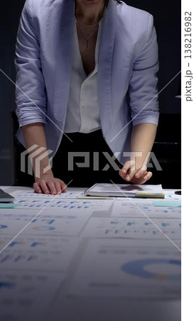 Businesswoman analyzing corporate financial data and sales growth charts on documents spread across an office desk, working late night in a professional setting Businesswoman analyzing corporate financial data and sales growth charts on documents spread across an office desk, working late night in a professional setting 138216982