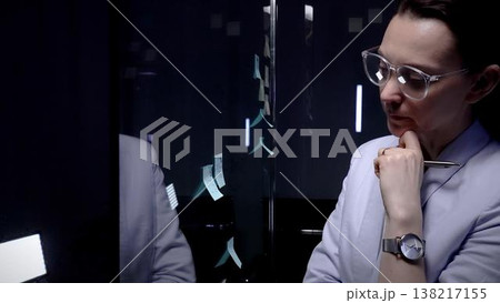 Professional businesswoman in glasses writing sticky notes on a glass planning board, brainstorming and organizing ideas for projects and strategy in a modern office setting 138217155