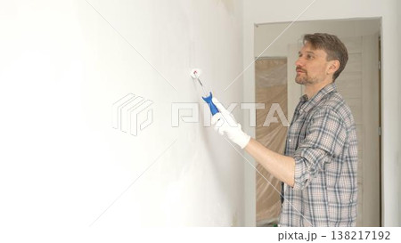 Man professional painter wearing checkered shirt, is rolling white paint on apartment wall, transforming interior space with steady strokes during comprehensive home renovation Man professional painter wearing checkered shirt, is rolling white paint on apartment wall, transforming interior space with steady strokes during comprehensive home renovation 138217192